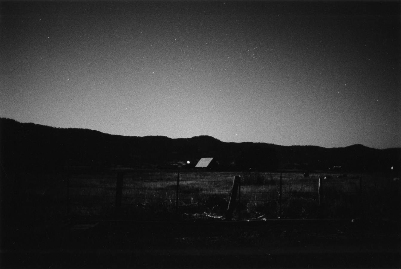 Black-and-white photograph of a vallery at dusk with a fence and a structure in the center fo the frame and mountains on the horizon