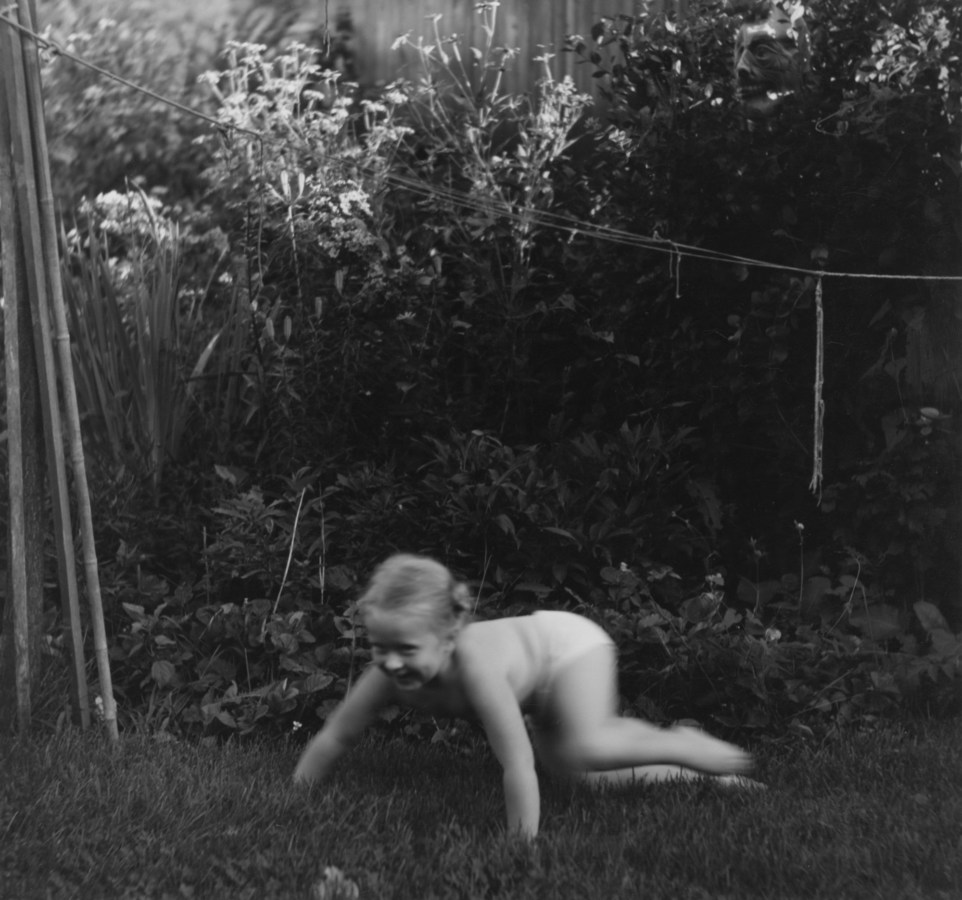 Black-and-white photograph of a young girl crawling through grass in a garden
