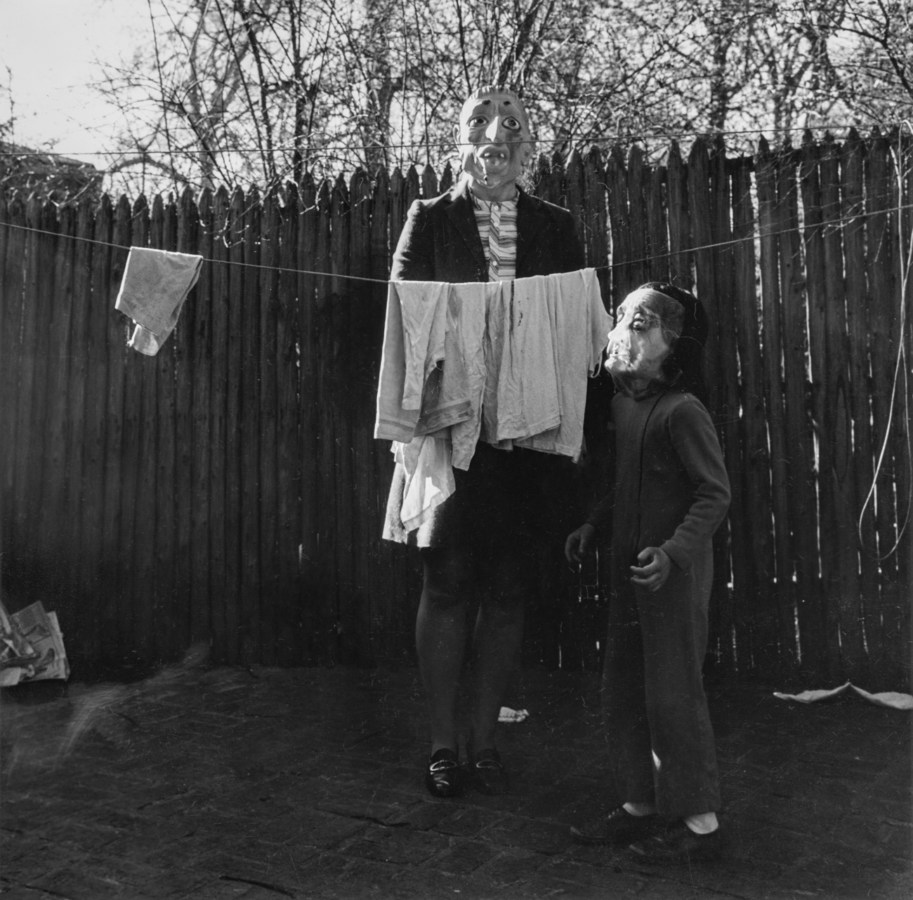 Black-and-white photograph of an adult and child in rubber masks standing behind a clothesline with a few garments