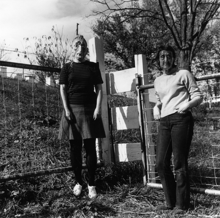Black-and-white photograph of two people in rubber masks standing by an ajar wooden gate