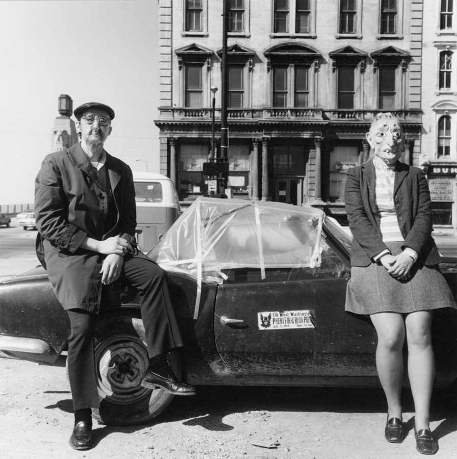 Black-and-white photograph of two people in rubber masks seated on a low convertible car