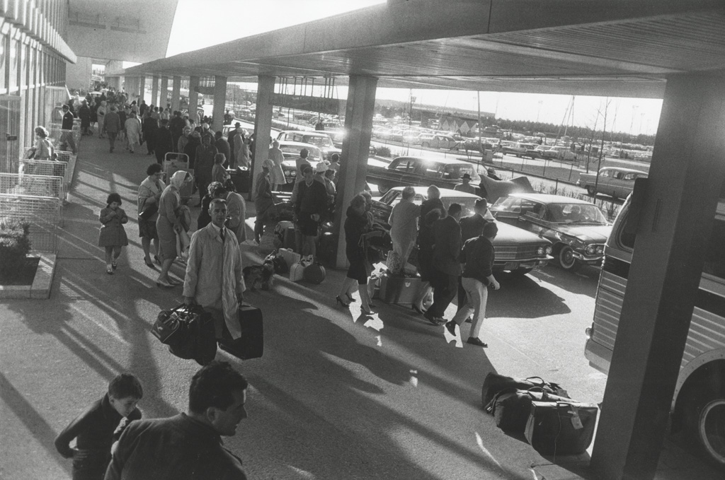 Black-and-white photograph of a sidewalk with passengers and luggage next to a narrow road with two lanes of cars