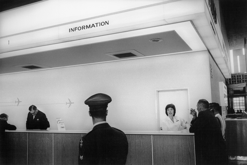 Black-and-white photograph of a pilot passing in front of an information desk staffed by a man and a woman