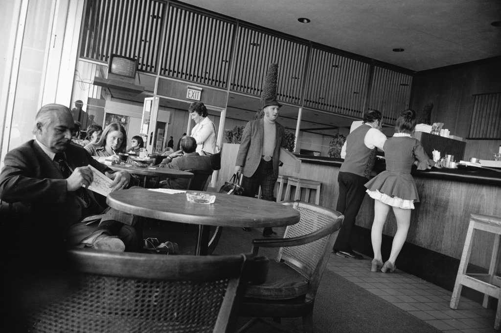 Black-and-white photograph of a man passing through a cafe area surrounded by tables and a counter