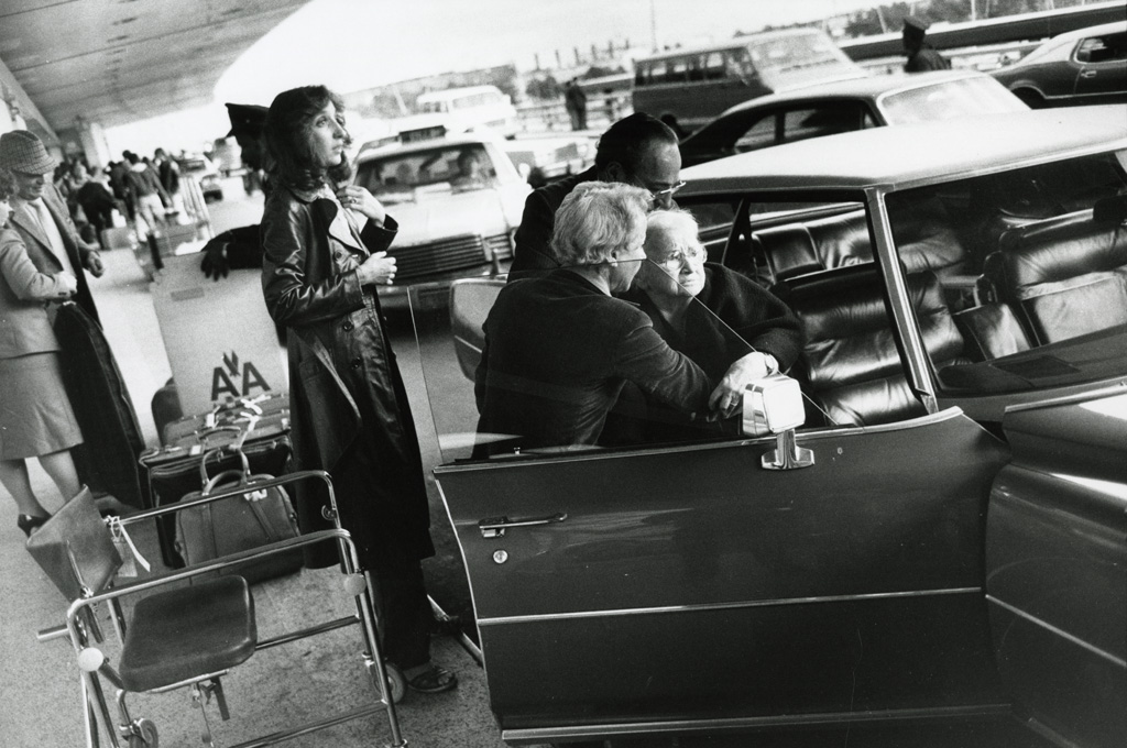 Black-and-white photograph of an elderly woman being helped into the passenger seat of a parked car