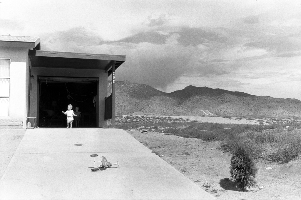 Black-and-white photograph of a toddler emerging from an open garage door against a mountainous desert landscape
