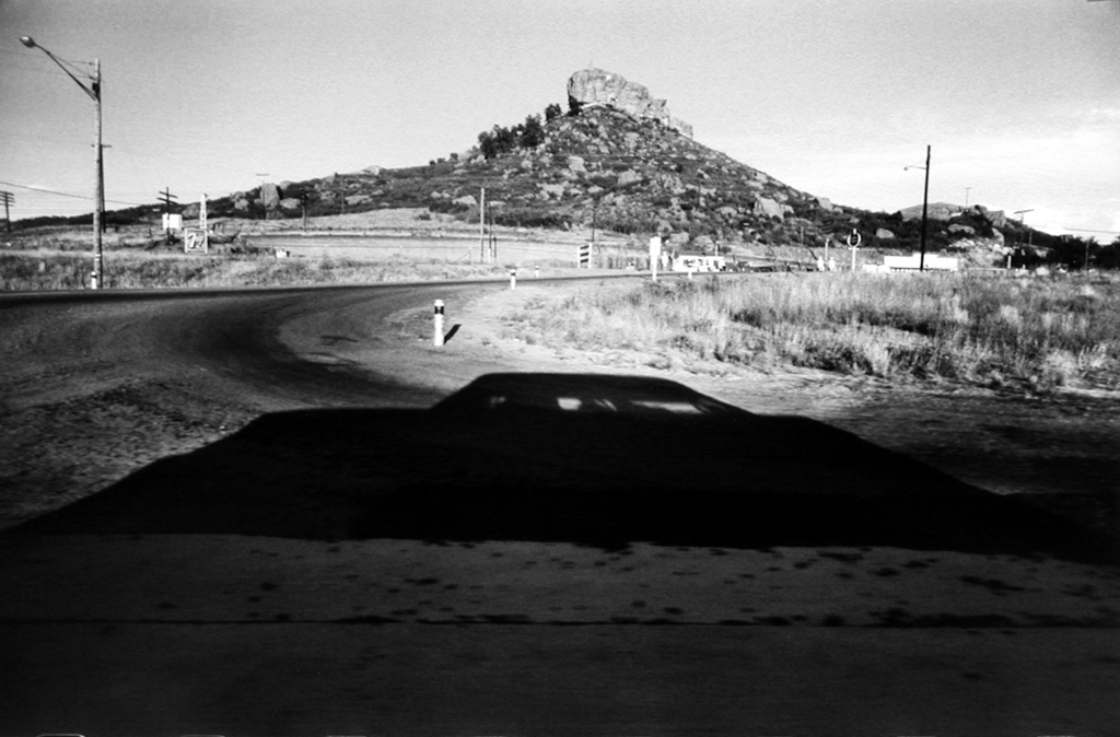 Black-and-white photograph of a roadside overlooking a rocky outcrop on a hill