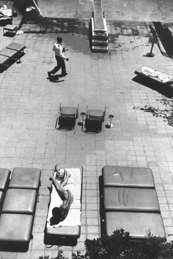 Black-and-white photograph taken from above of a man crossing a pool deck in front of a woman in a lounge chair