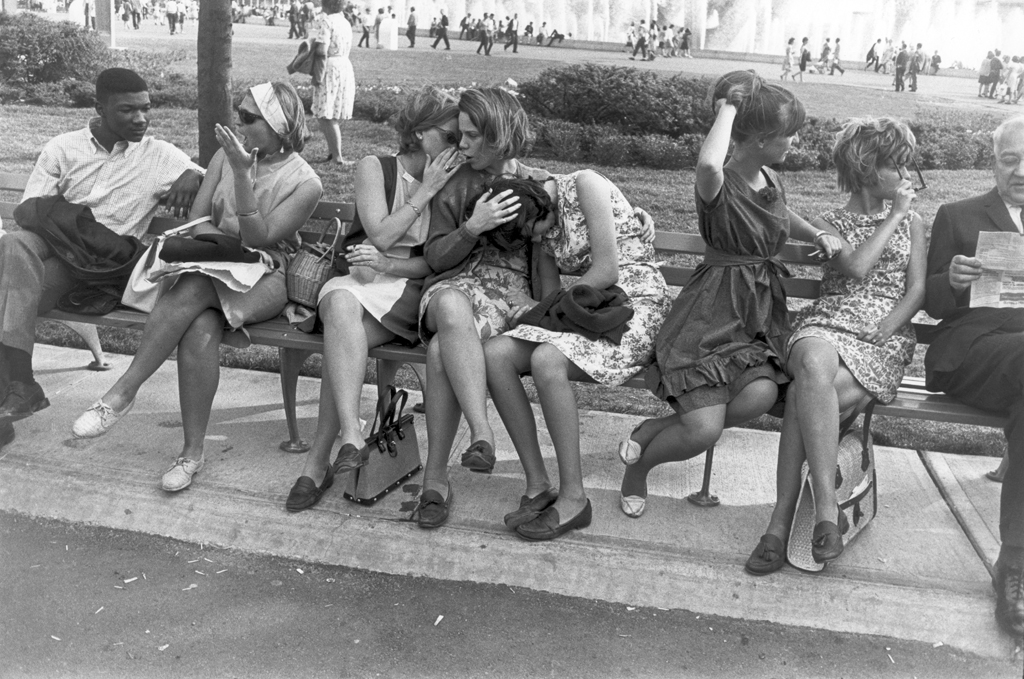 Black-and-white photograph of six young women on a park bench bordered by a young man conversing and an older man reading a newspaper