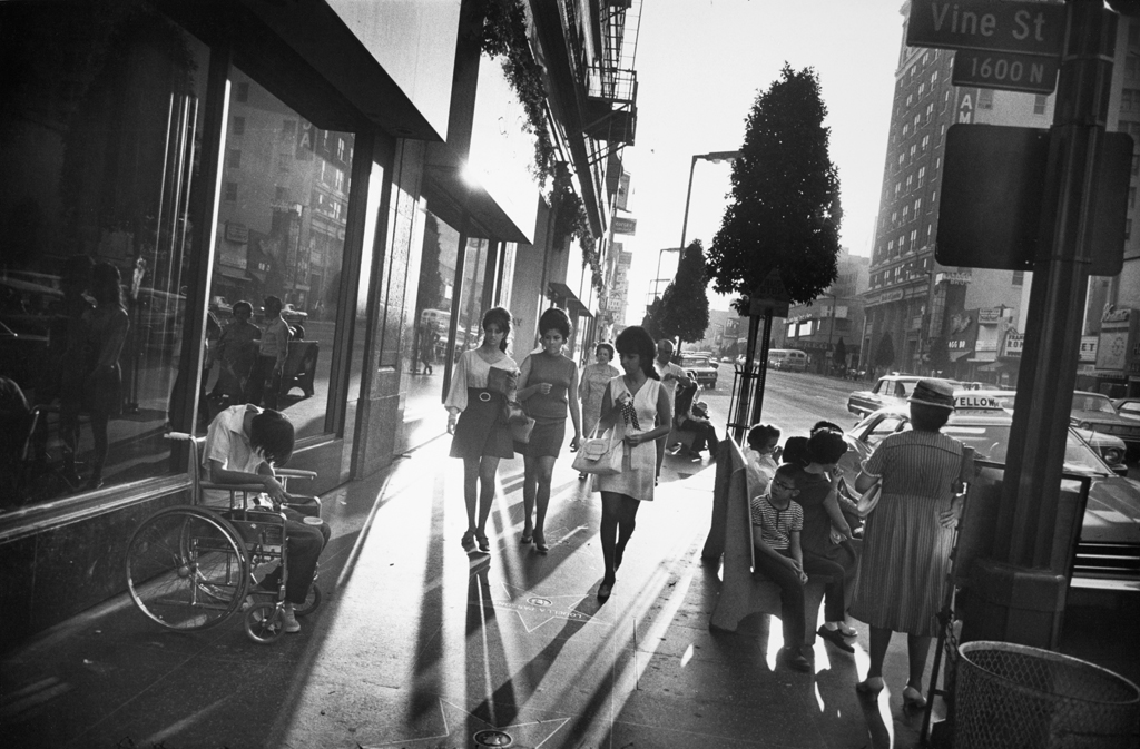 Black-and-white photograph of three women walking down a city sidewalk between a person in a wheelchair and a crowded bench