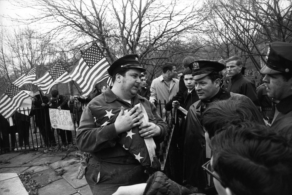 Black-and-white photograph of a man holding a folded American flag conversing with people across a fence