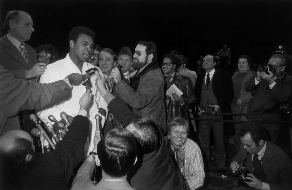 Black-and-white photograph of a boxer being interviewed by reporters with microphones and cameras