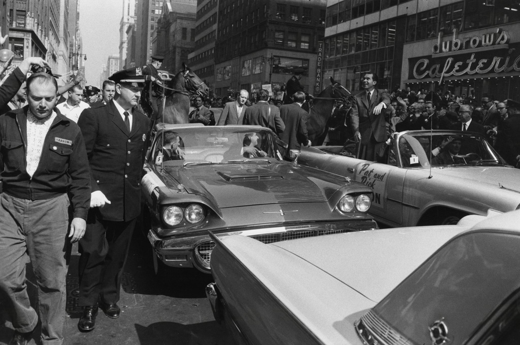 Black-and-white photograph of a parade of convertible cars moving down a crowded avenue
