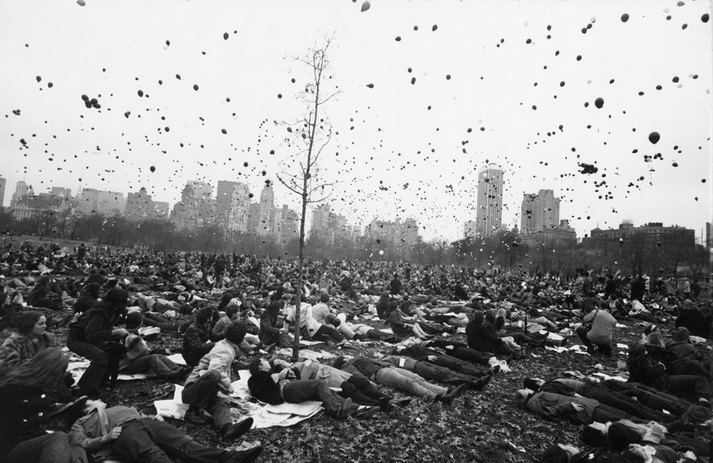 Black-and-white photograph of people sitting on blankets in a meadow under a sky filled with helium balloons