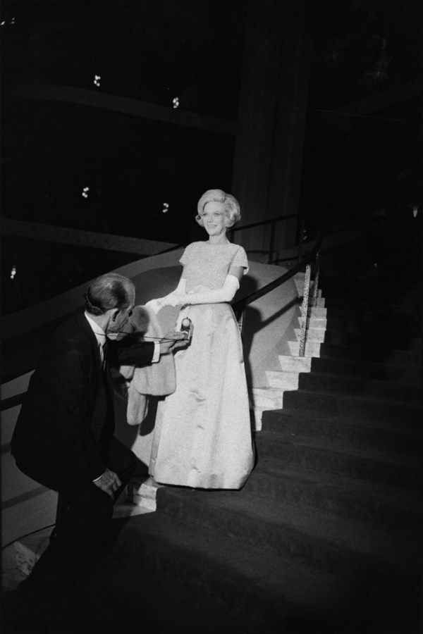 Black-and-white photograph of a woman in a pale gown descending a curving staircase towards a man in a suit