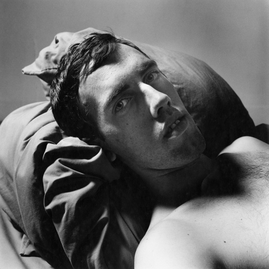 Black-and-white photograph of a shirtless man reclining on pillows