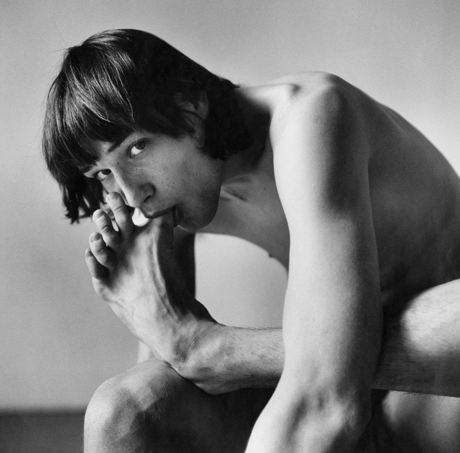 Black-and-white photograph of a seated nude man leaning forward to suck his big toe