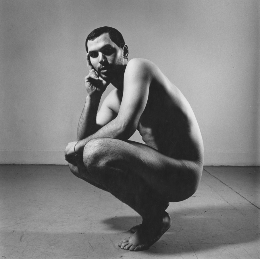 Black-and-white photograph of a crouching nude man with his cheek on one hand
