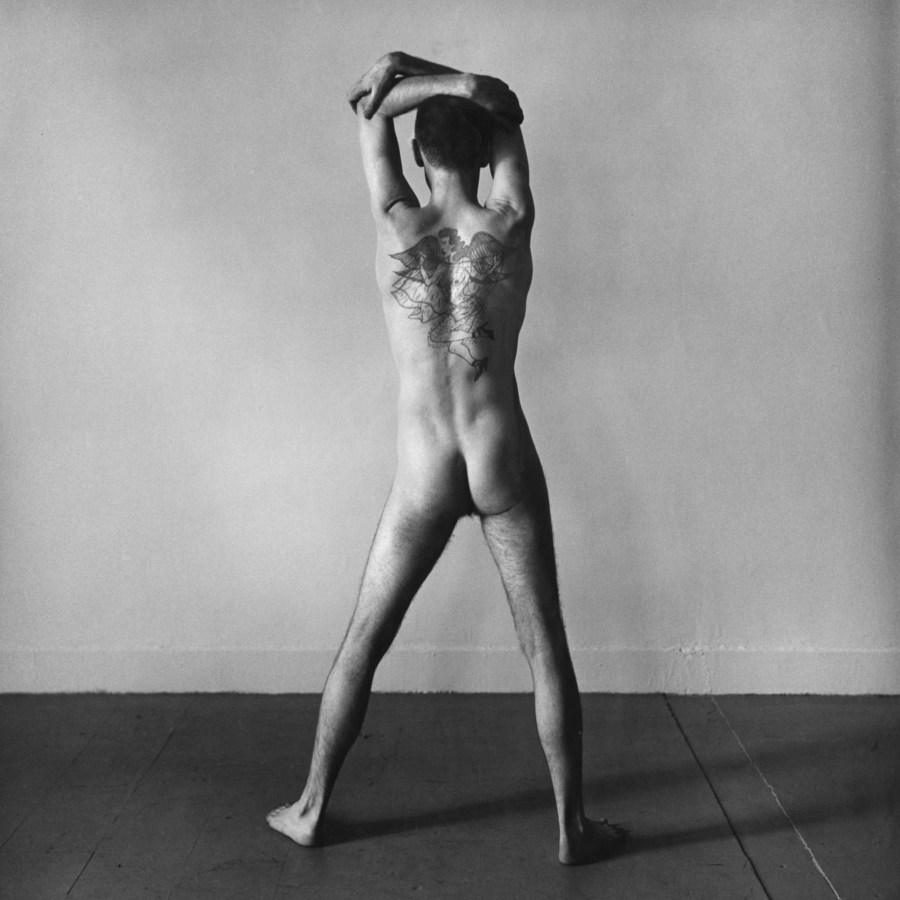 Black-and-white photograph of a standing nude man with his tattooed back to the viewer and his arms above his head