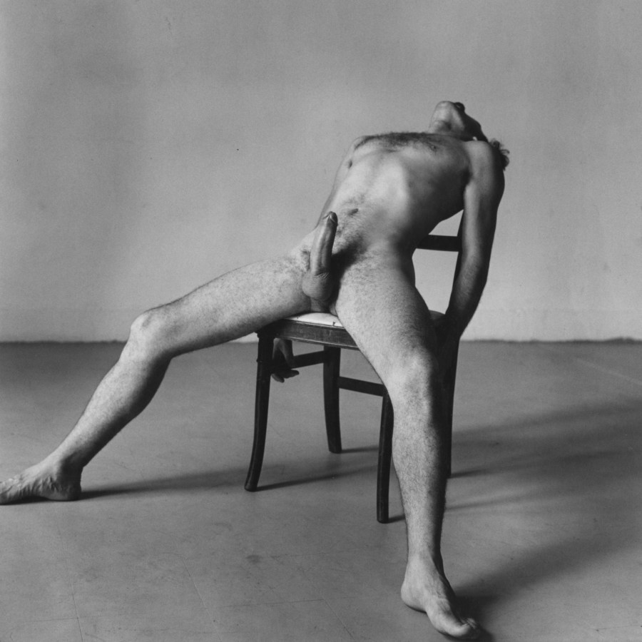 Black-and-white photograph of a seated nude man with an erection bending backward in his chair