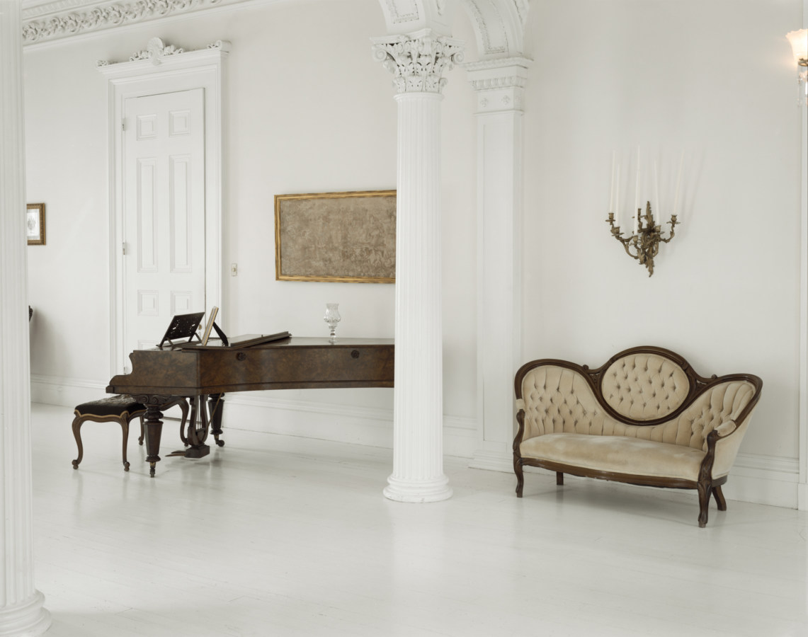 Color photograph of a white room with Neo-Classical detailing containing a beige sofa and dark wooden grand piano