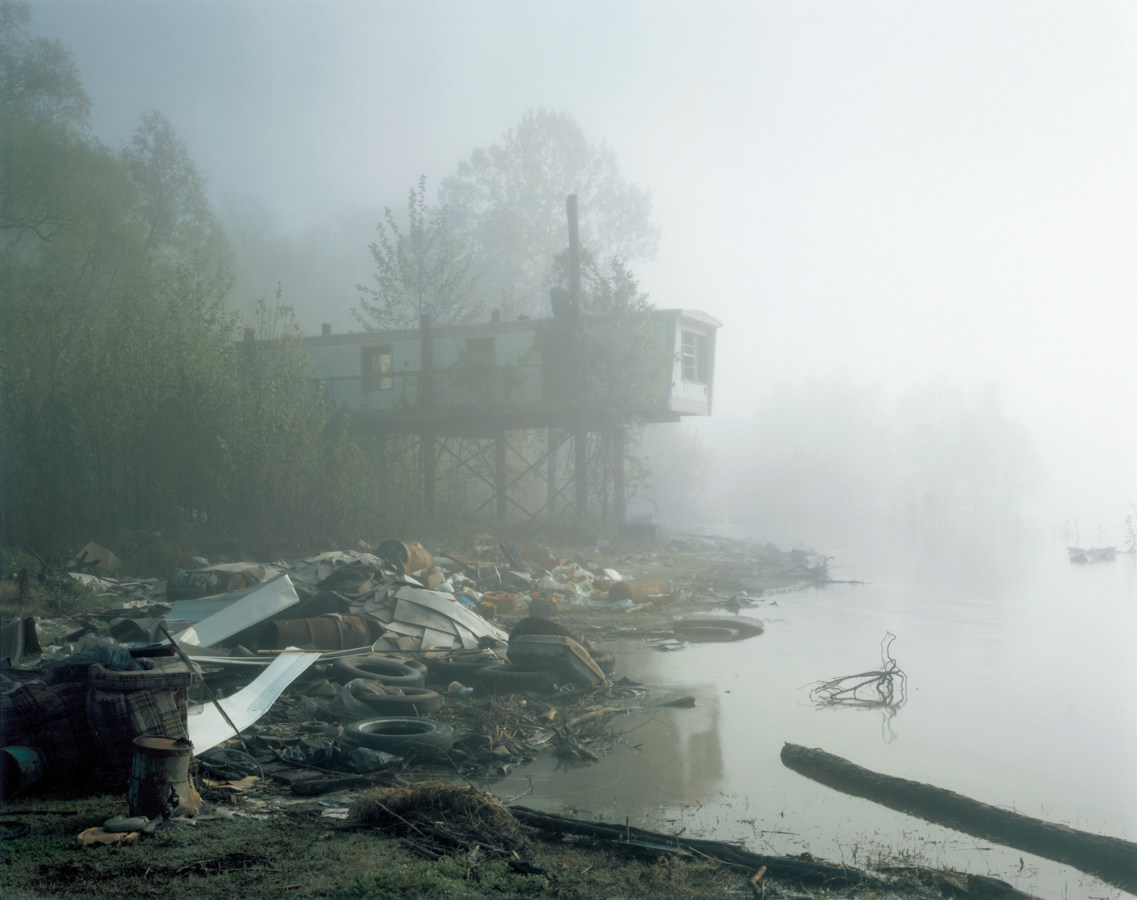 Color photograph of a decrepit trailer home on stilts standing above a shore filled with trash