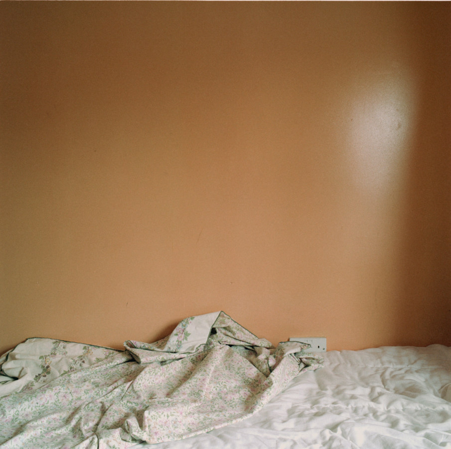 Color photograph of an umade bed in front of a burnt orange wall.