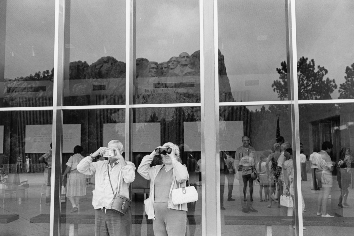 Black-and-white photograph of an elderly couple looking with binoculars towards the left. Mt. Rushmore is reflected in the windows behind them.