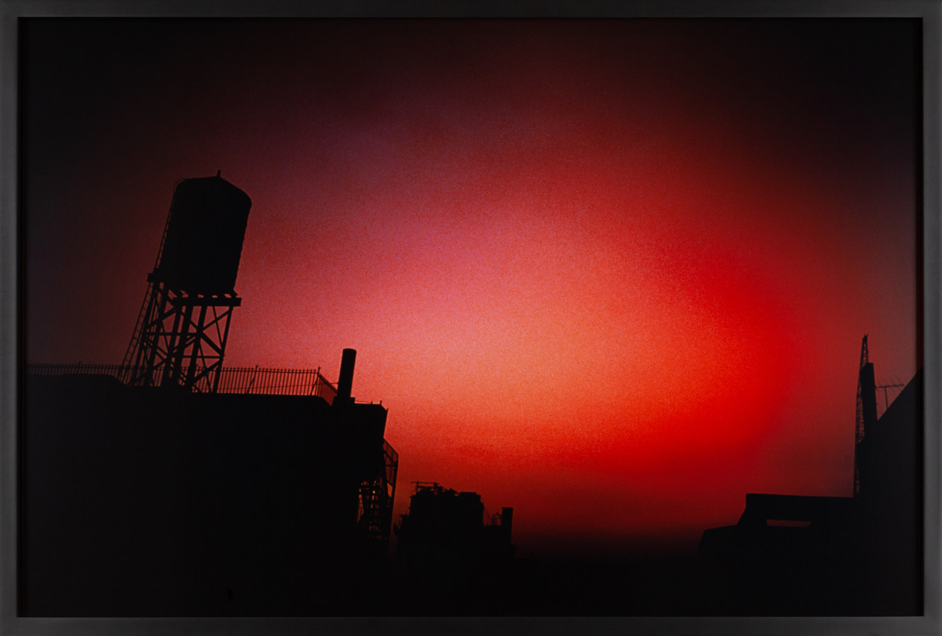 Color photograph of an overcast night sky with a red glow above city buildings and a rooftop watertower