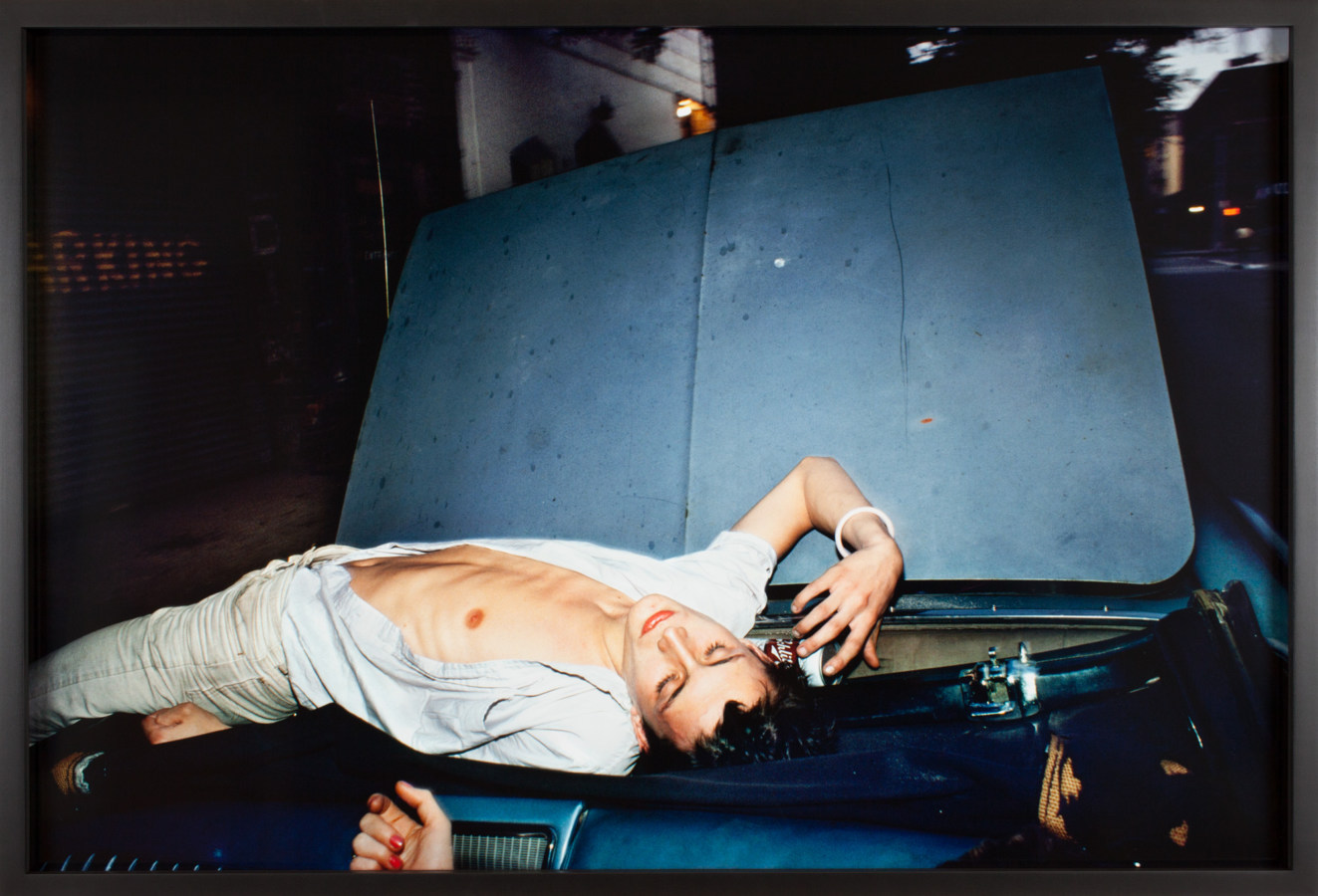 Color photograph of a young man with an open white shirt lying on the windshield of a blue car