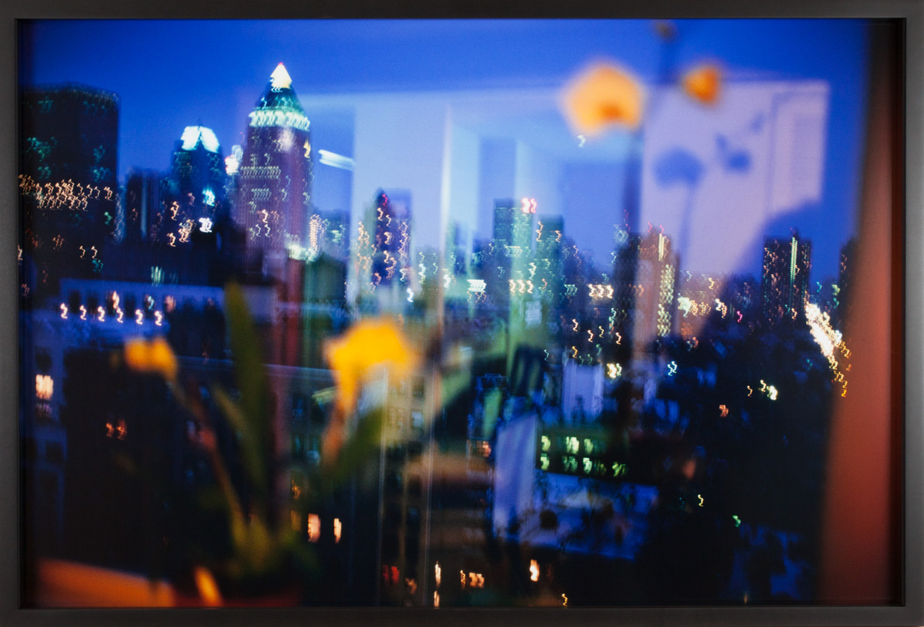Color photograph from behind a window of an out-of-focus city skyline at night with faint reflections from the room superimposed