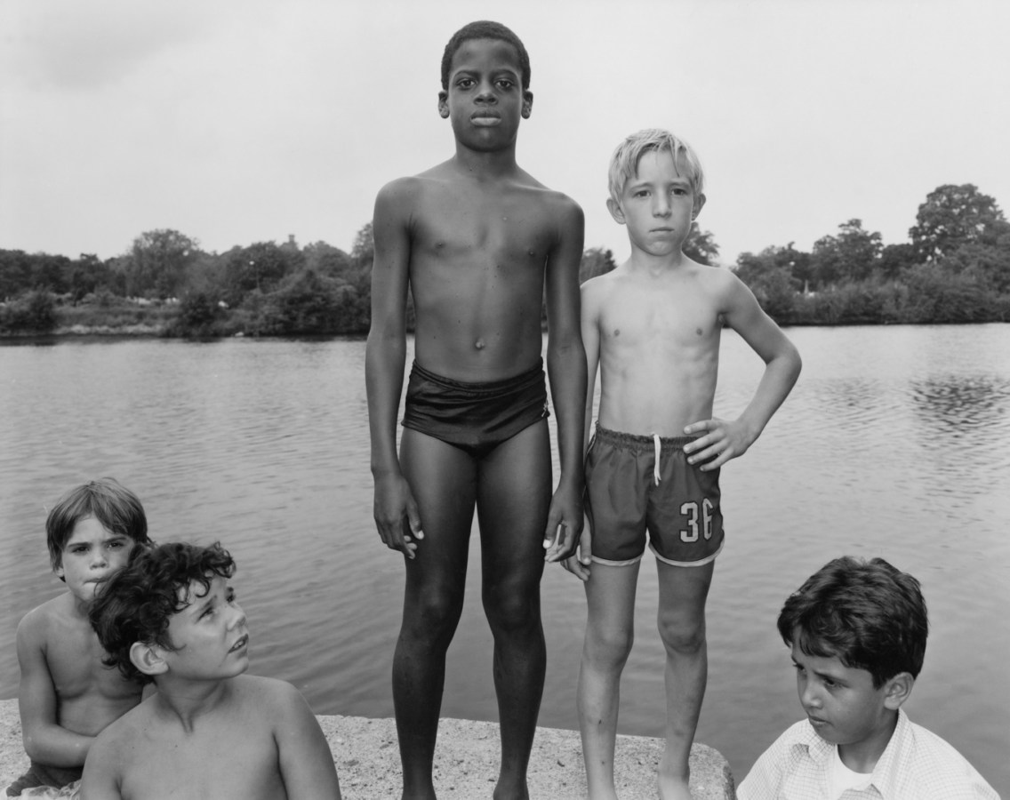 Black and white photograph of two boys in swimsuits standing on a concrete ledge above a lake with three others seated beneath them