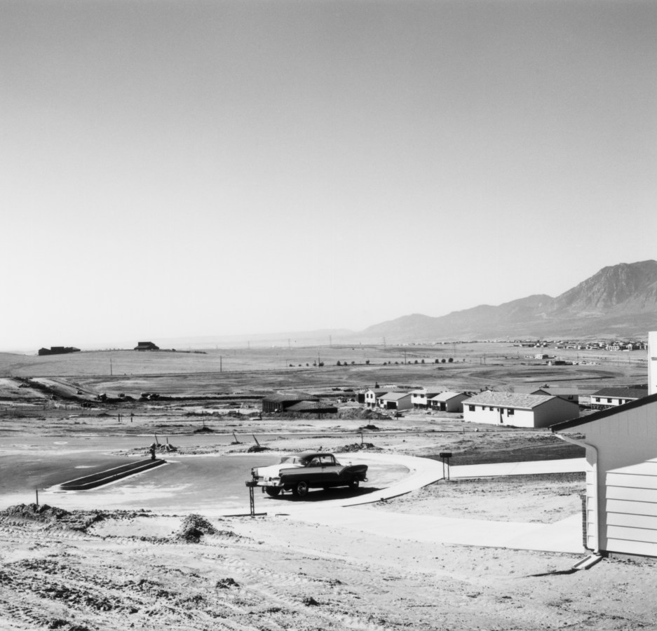 A black and white photograph of a lone car parked in a cul de sac, with houses and mountains in the background.