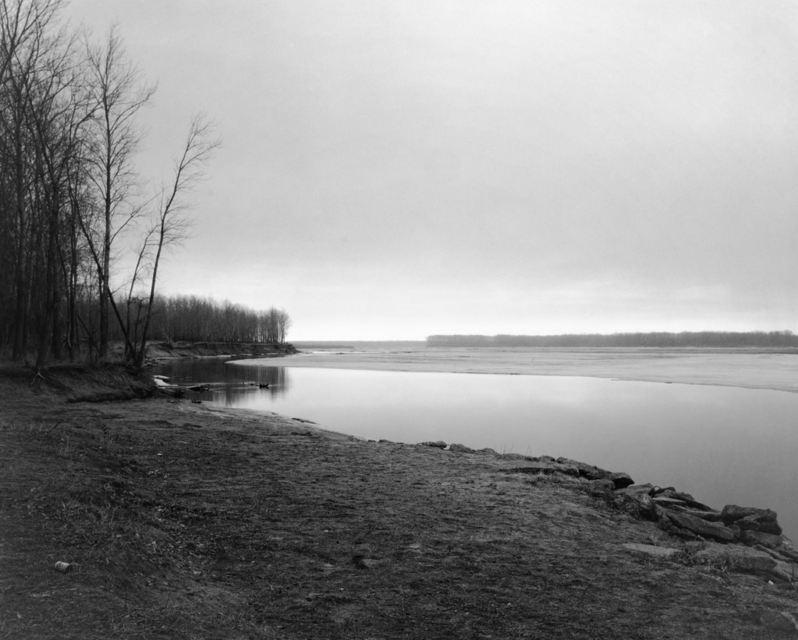 A black and white photograph of a river bank with leafless trees at the right edge of photograph and an overcast sky.
