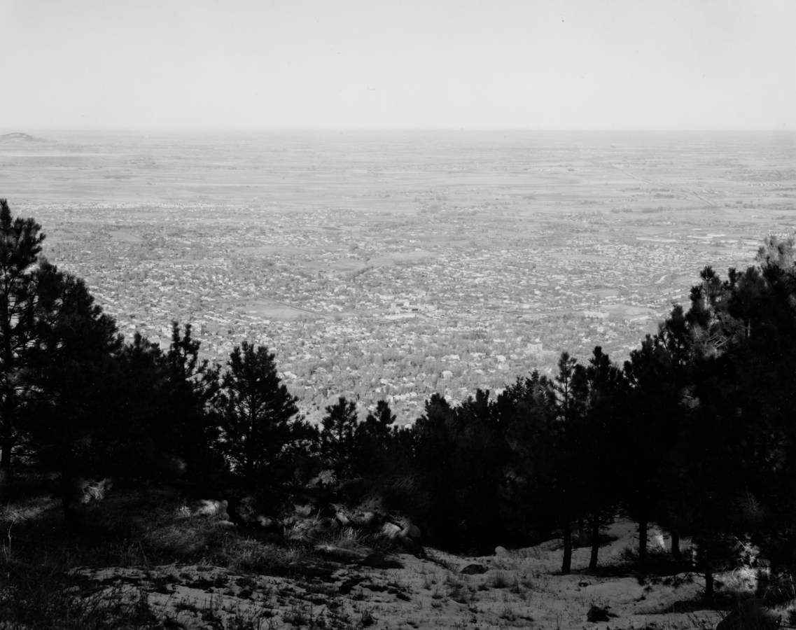 Black and white photograph of a tree-covered hillside overlooking a town on the flat plain below