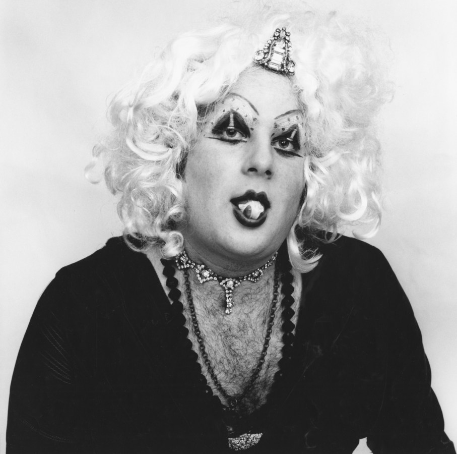 Black-and-white figure of a sitter wearing a blonde wig, heavy makeup, and a deep-cut shirt showing off a hairy chest, popping chewing gum