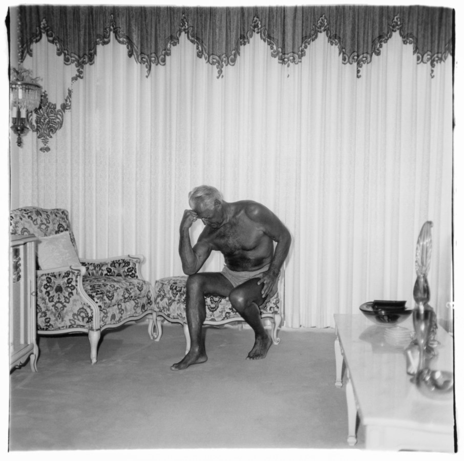 Black and white photograph of a man flexing his arm muscles seated a living room