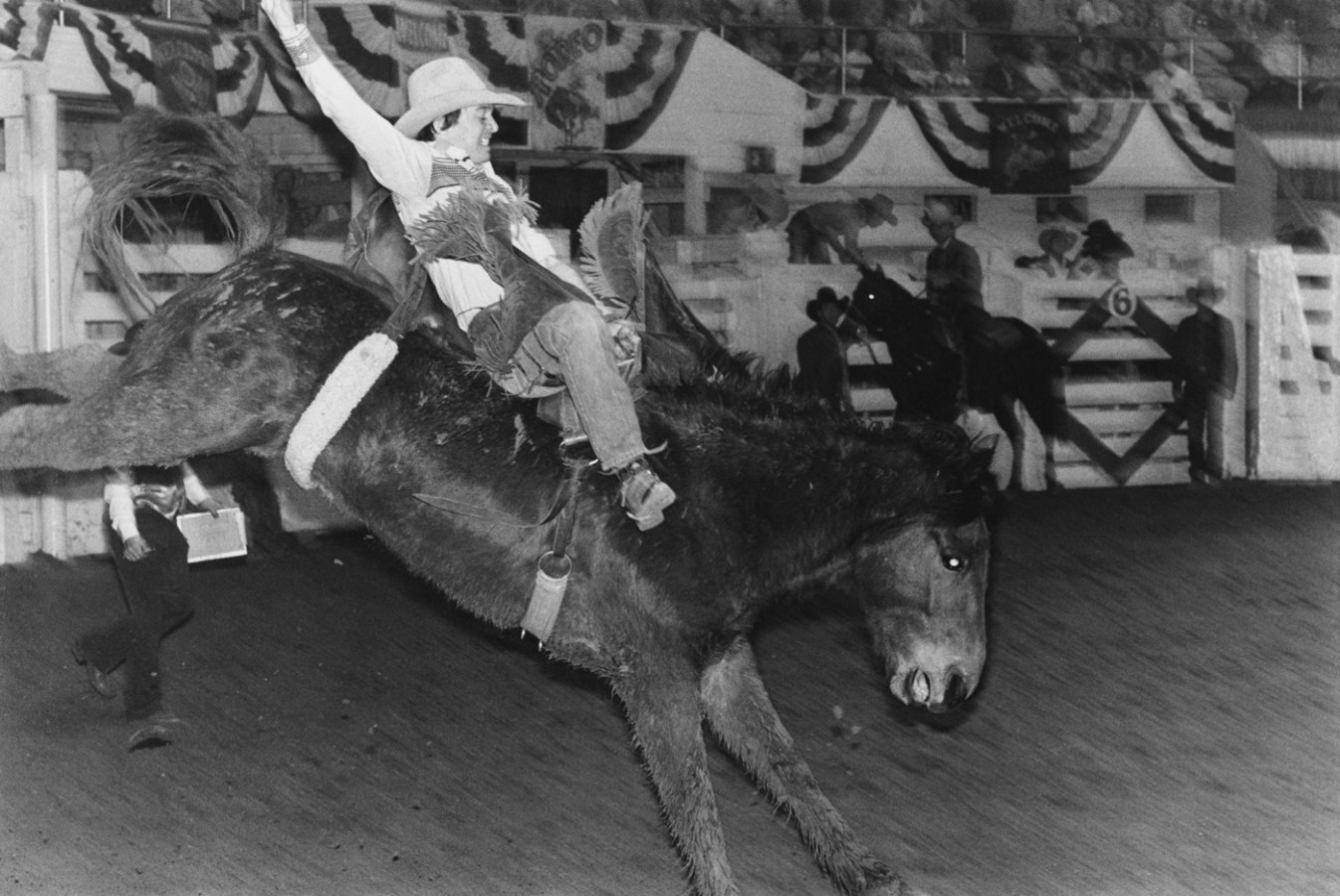 Black-and-white photograph of a cowboy with one arm raised sitting astride a bucking horse