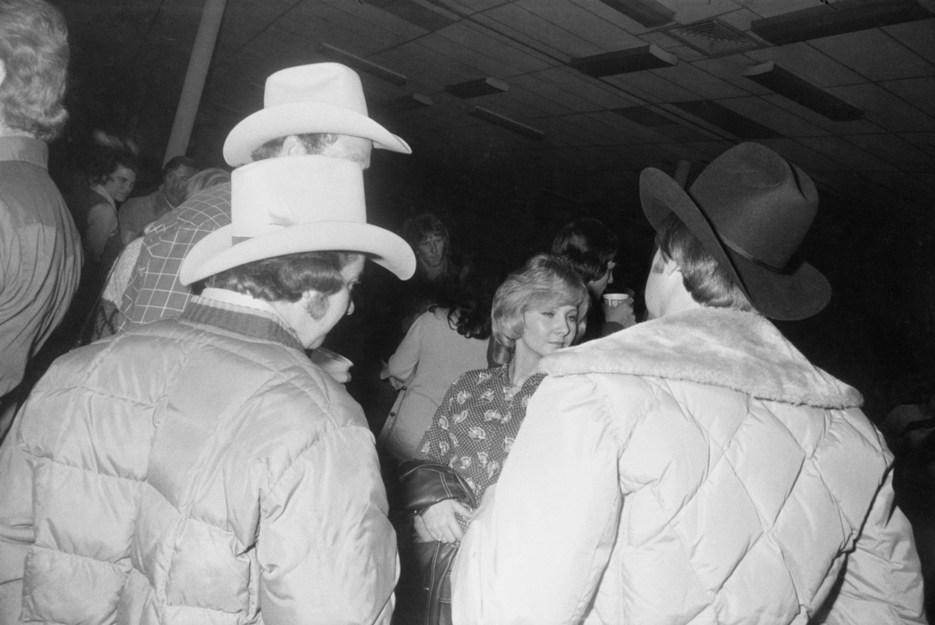 Black-and-white photograph of men in cowboy hats with their back to the viewer conversing with a woman