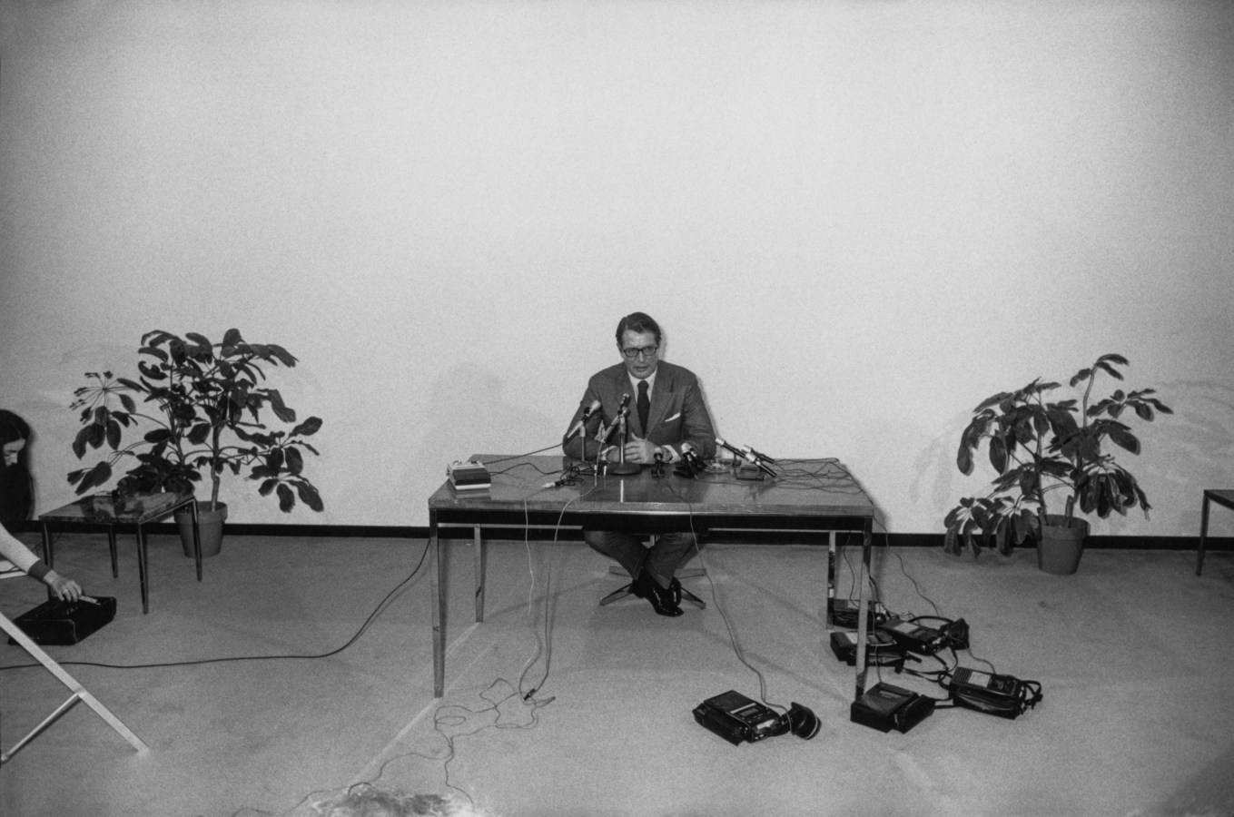 Black-and-white photograph of a man behind multiple microphones at a table against a blank wall