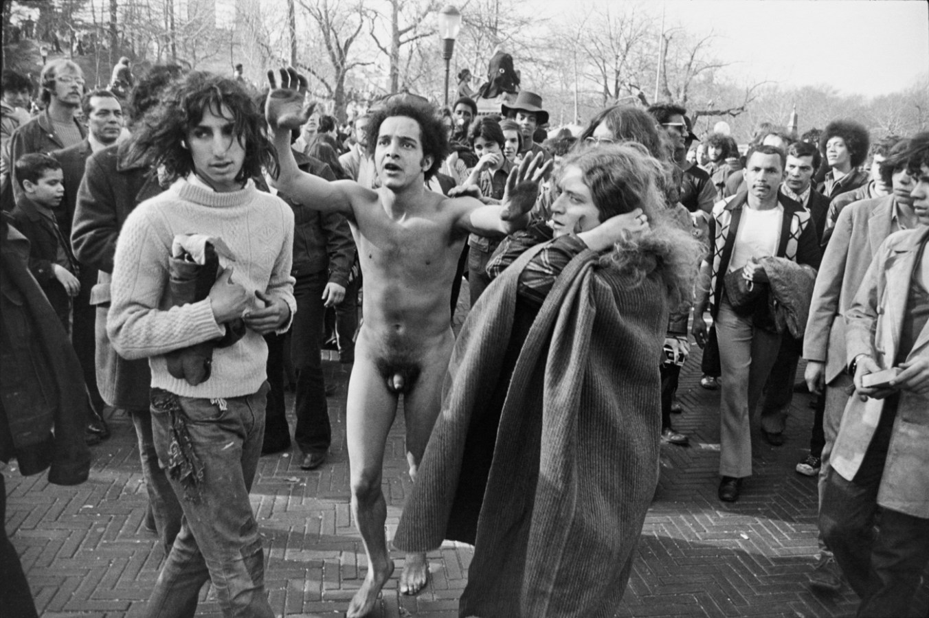 Black-and-white photograph of a naked man with raised arms in a crowded park
