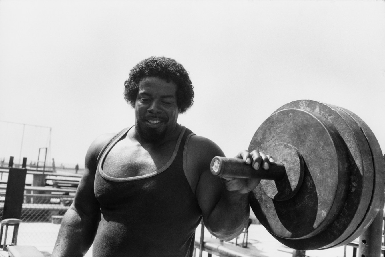 Black-and-white photograph of a muscular man in a tank top next to a large barbell weight