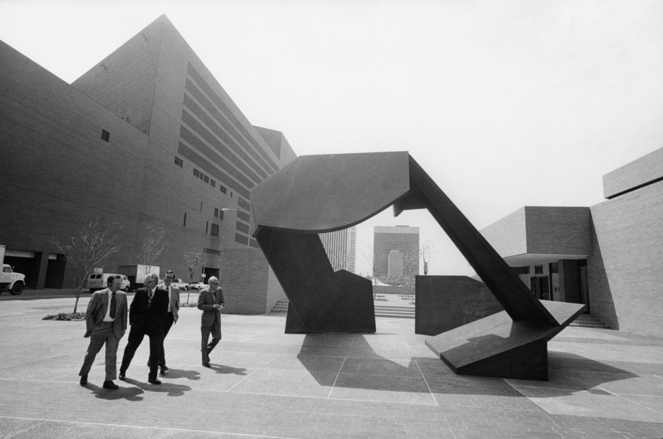 Black-and-white photograph of a group of businessmen passing an empty plaza with a modern geometric sculpture