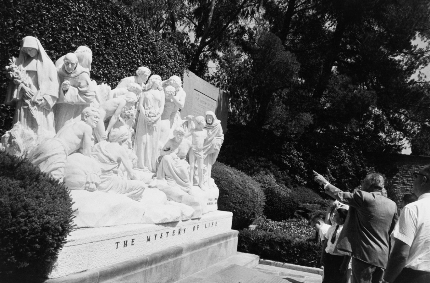 Black-and-white photograph of a man pointing out a large group memorial sculpture titled THE MYSTERY OF LIFE to a small group