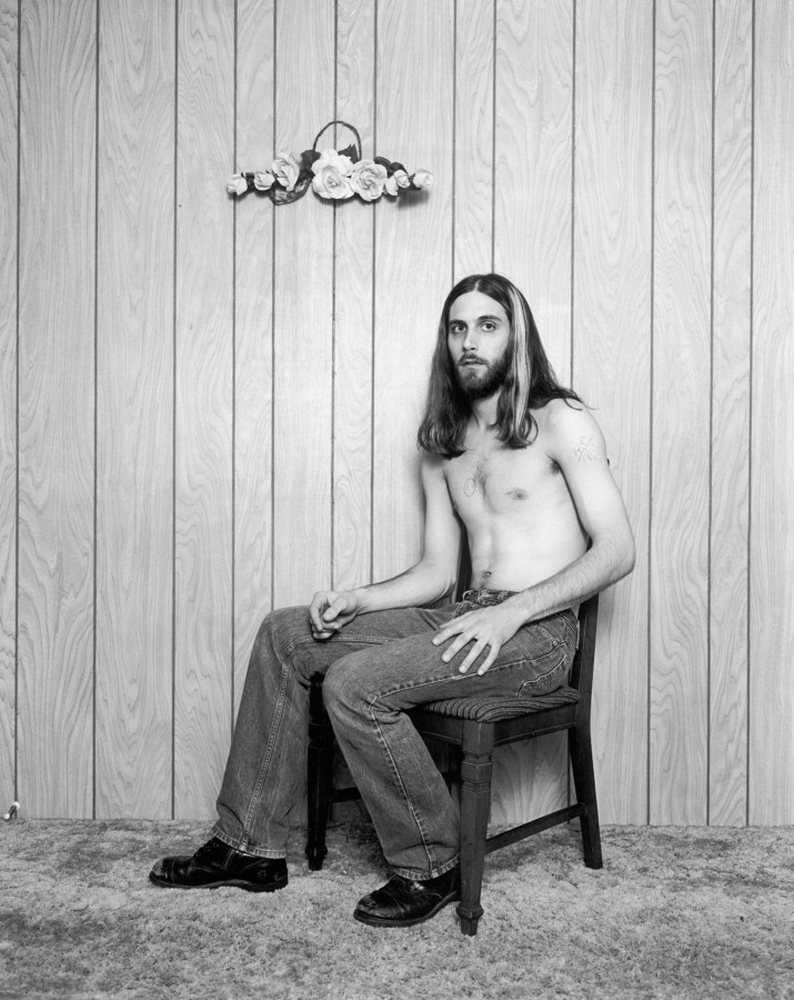 Black-and-white photograph of a shirtless man with long hair seated in front of a wood paneled wall
