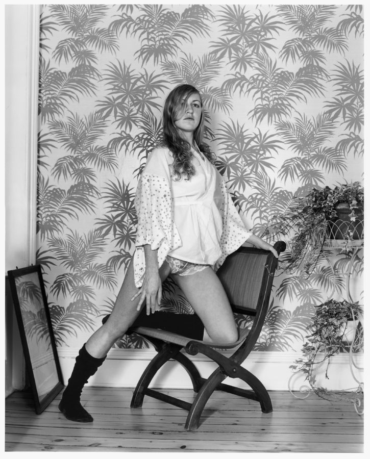 Black-and-white photograph of a woman resting one knee on a chair in front of a palm-leaf patterned wallpaper
