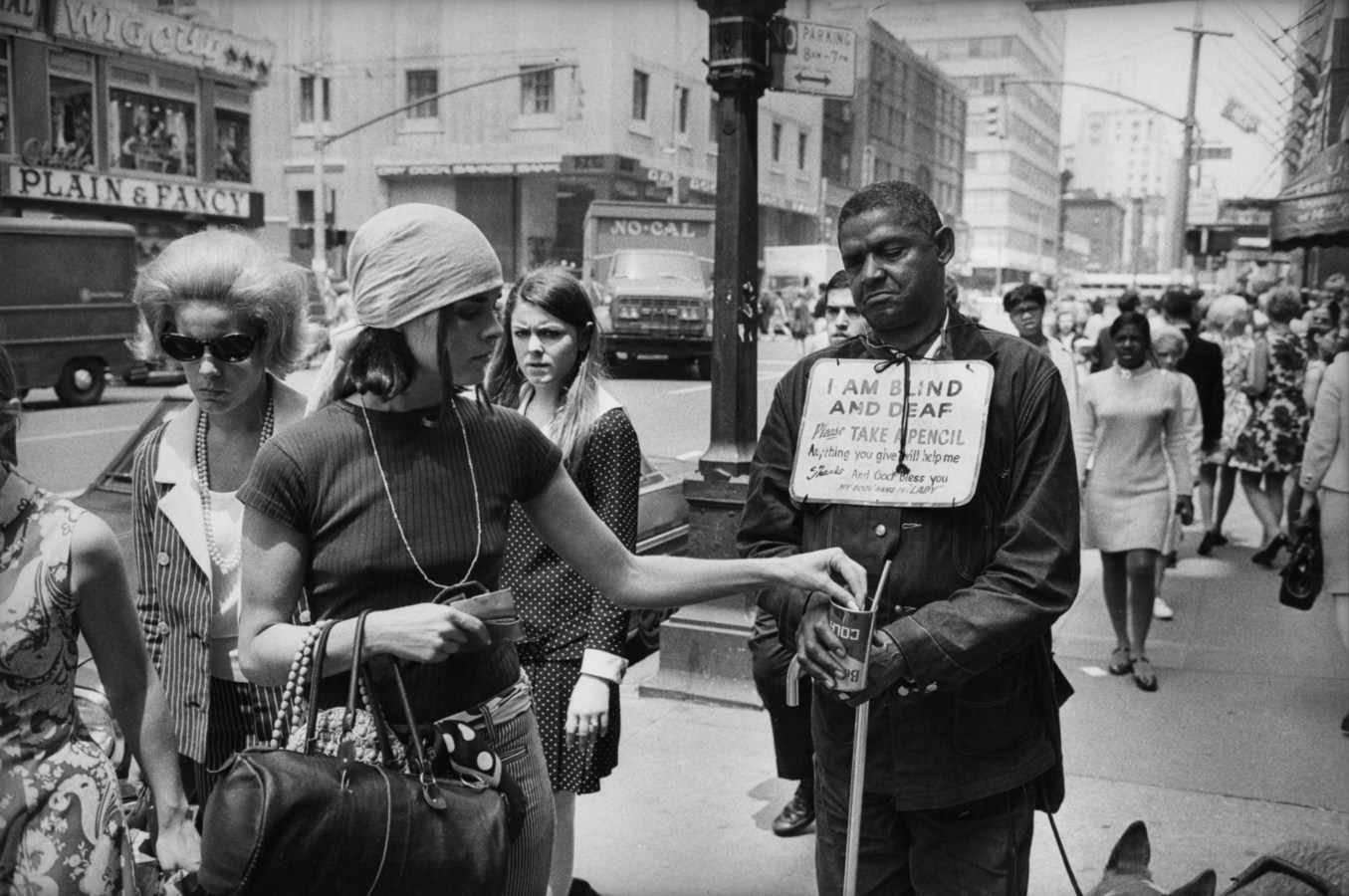 Black and white photograph of a bustling street scene depicting a women donating money to a person on street with sign