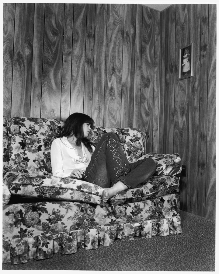 Black-and-white photograph of a woman seated on a flowered sofa in a wood paneled room