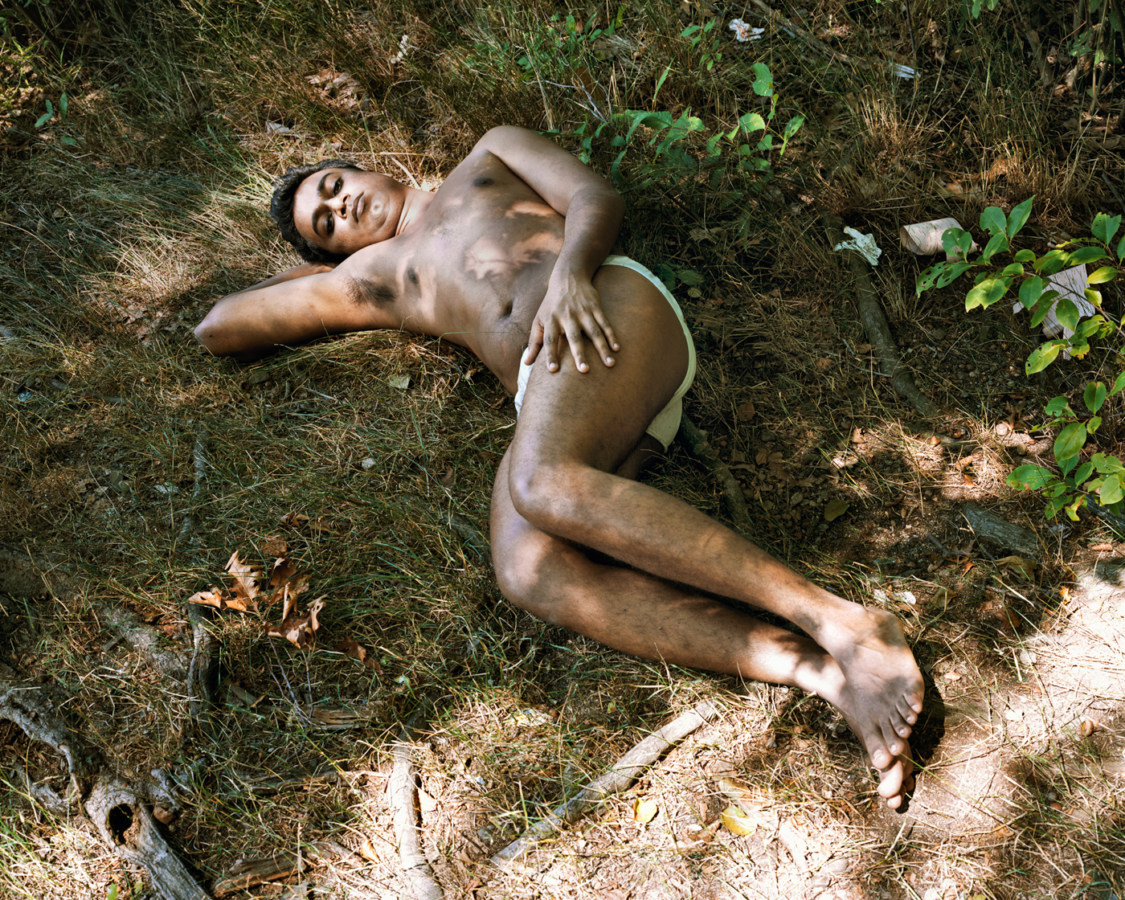 Color photograph of a shirtless man reclining on his side in sparse grass
