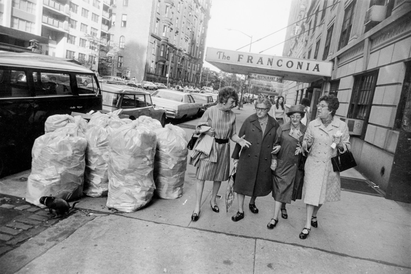 Black-and-white photograph of a group of older women conversing and walking past a pile of trash bags on the sidewalk